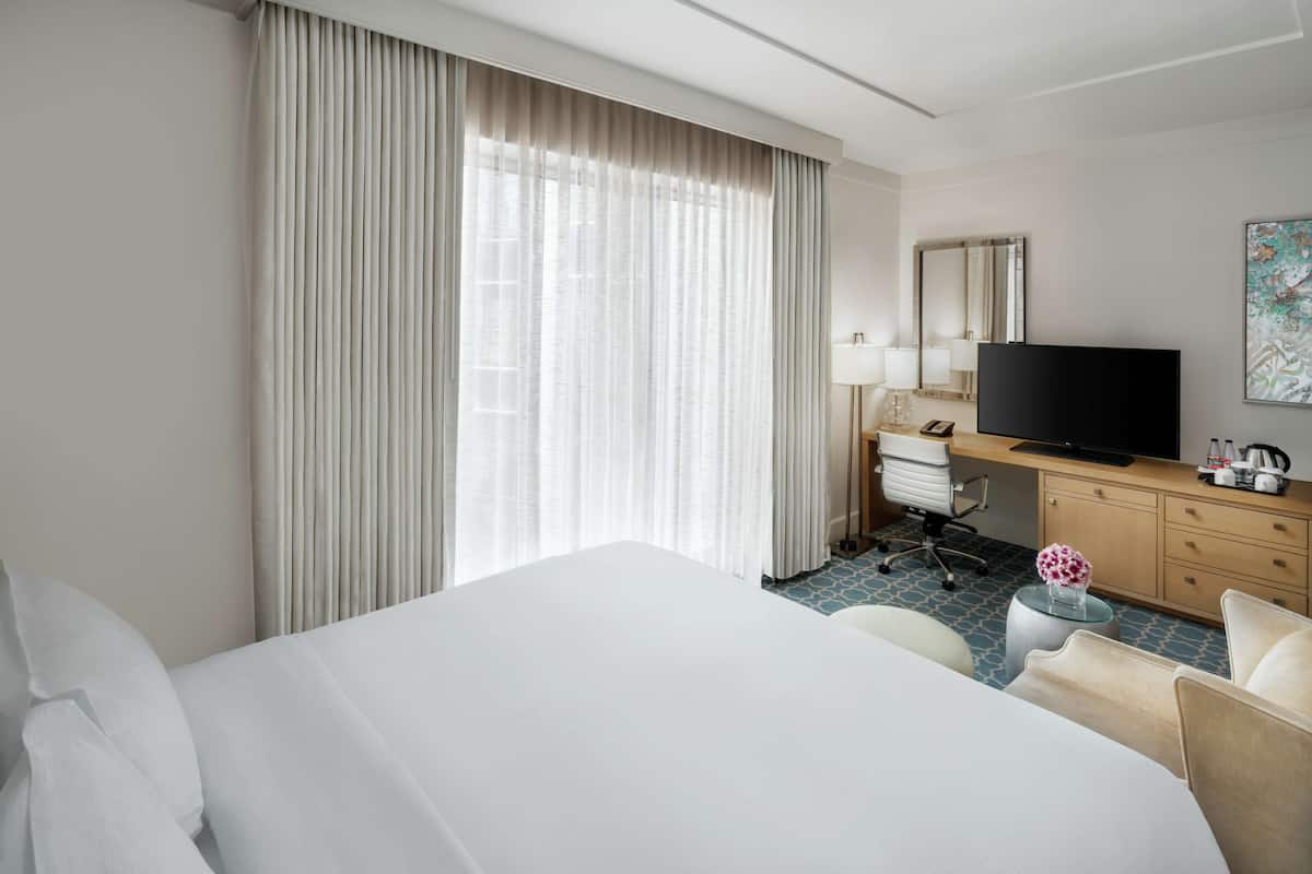A hotel room with a large bed, a desk with a television, a chair, and a small table with a vase.