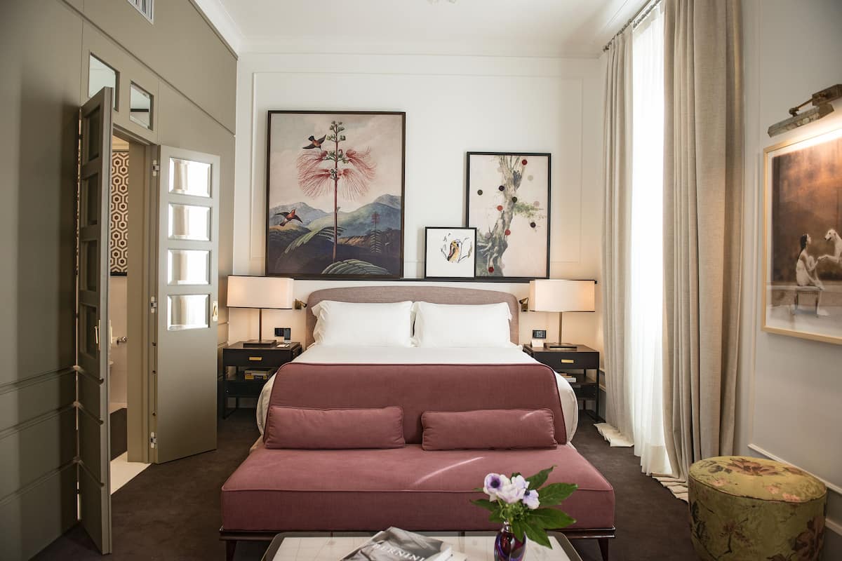 A hotel room with a large bed, two bedside tables with lamps, a desk, and a chair. There are three framed artworks on the wall.