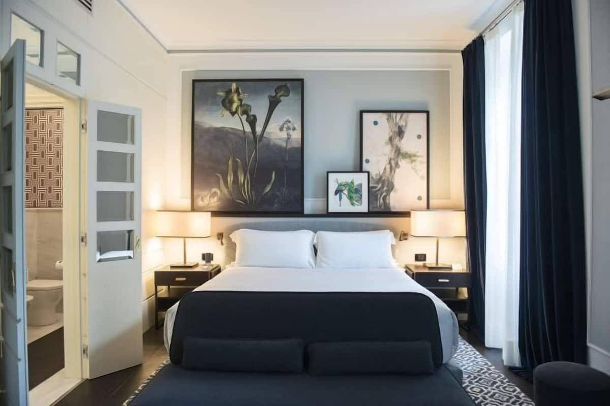 A hotel room with a bed, two bedside tables, a nightstand, a door leading to a bathroom, and two wall-mounted artworks.