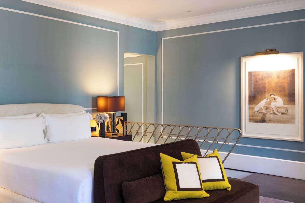 A hotel room with a bed, a brown sofa with cushions, a bedside table with a lamp, and a framed picture on the wall.