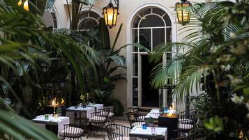 An indoor dining area with tables and chairs, surrounded by lush tropical plants and arched windows.
