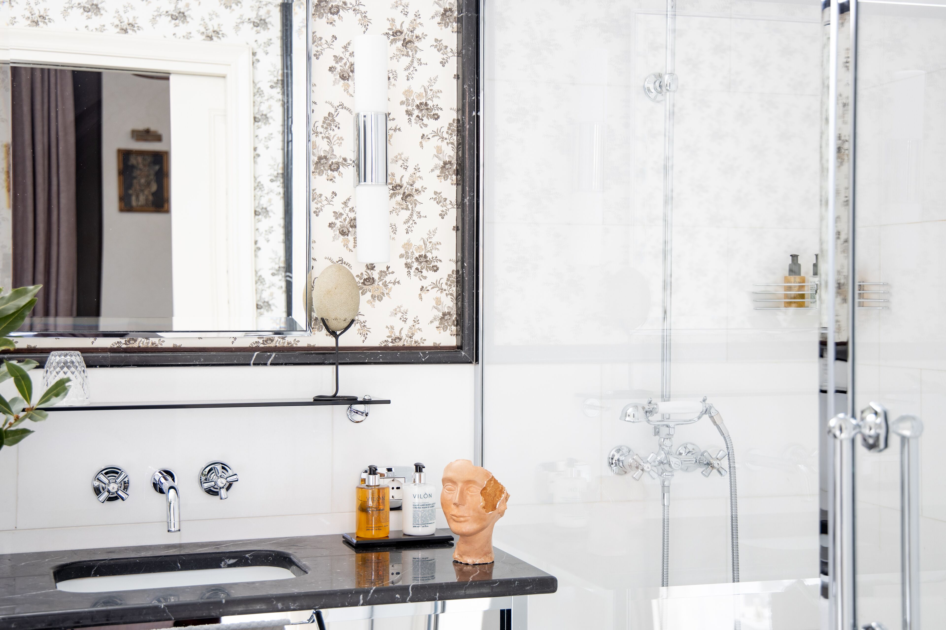 Une salle de bain avec un lavabo en marbre, une douche et un miroir orné d’un papier peint à motifs floraux.