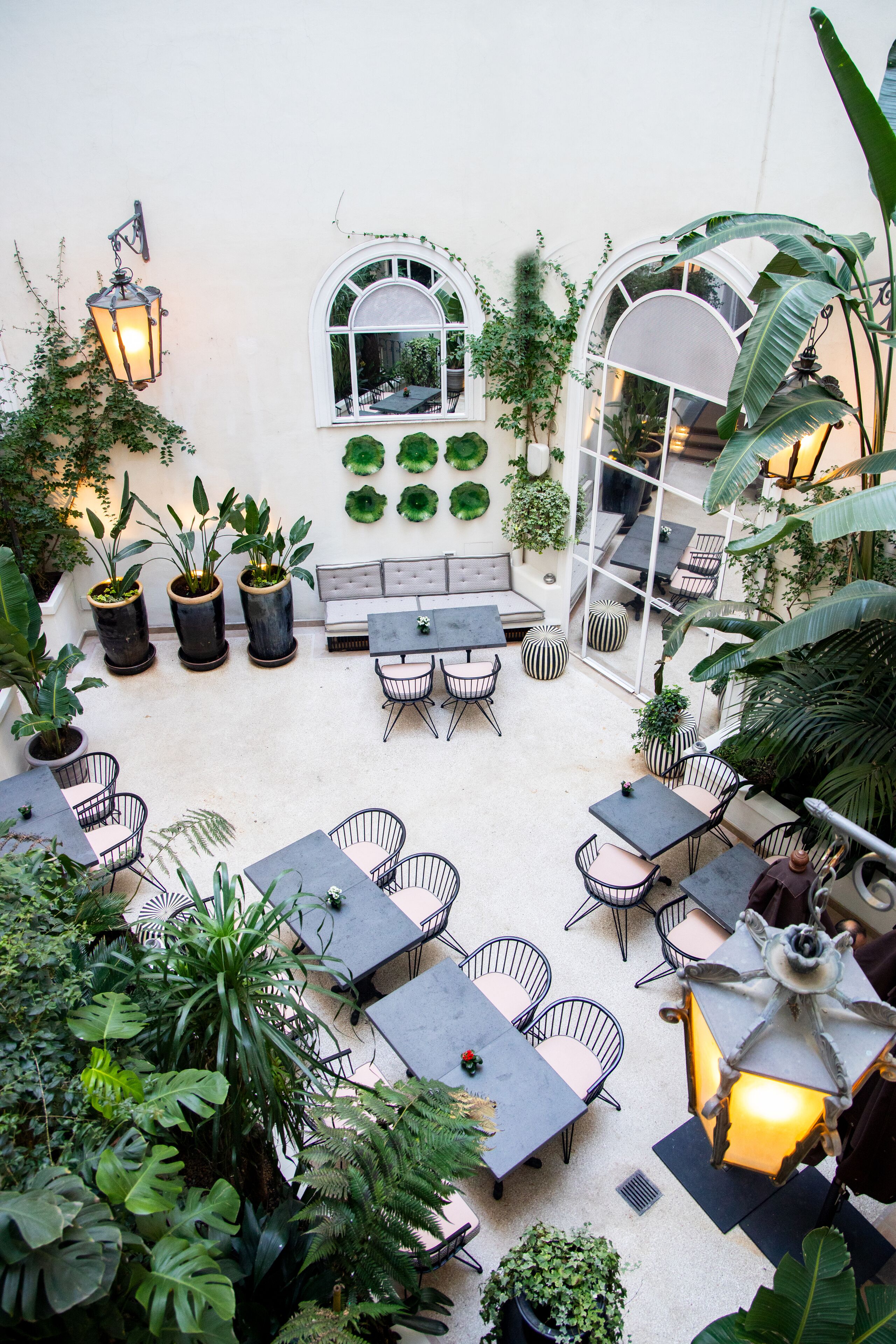 Une terrasse extérieure avec des tables et des chaises, entourée de plantes en pot et d’un mur blanc percé de fenêtres.