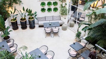 Une terrasse extérieure avec des tables et des chaises, entourée de plantes en pot et d’un mur blanc percé de fenêtres.
