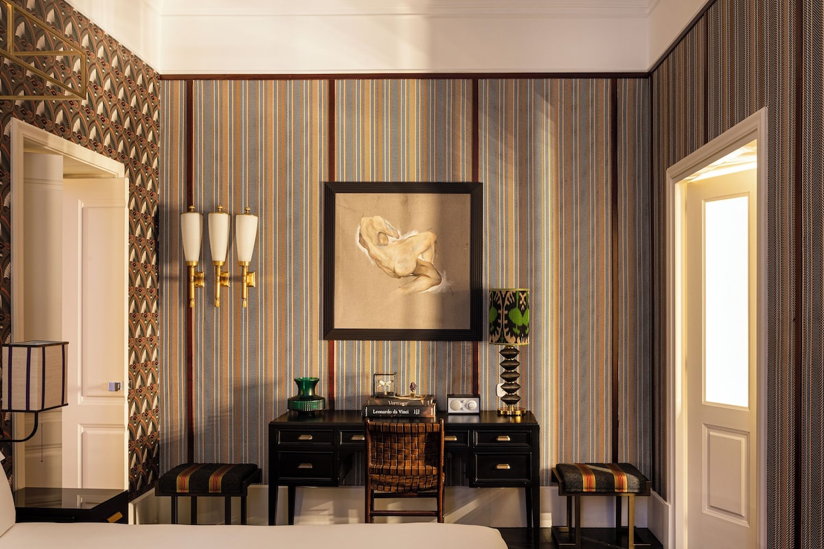 A hotel room with a large bed, a desk with drawers, two chairs, a framed artwork on the wall, and a patterned wallpaper.