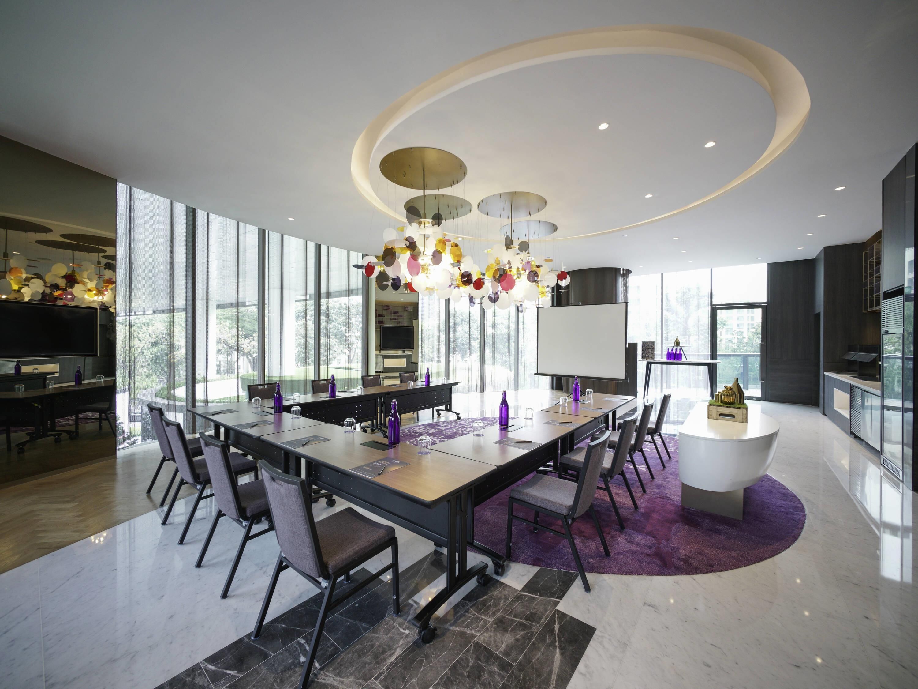 A modern dining area with a long table, chairs, a central chandelier, and a large window offering a view of the outdoors.