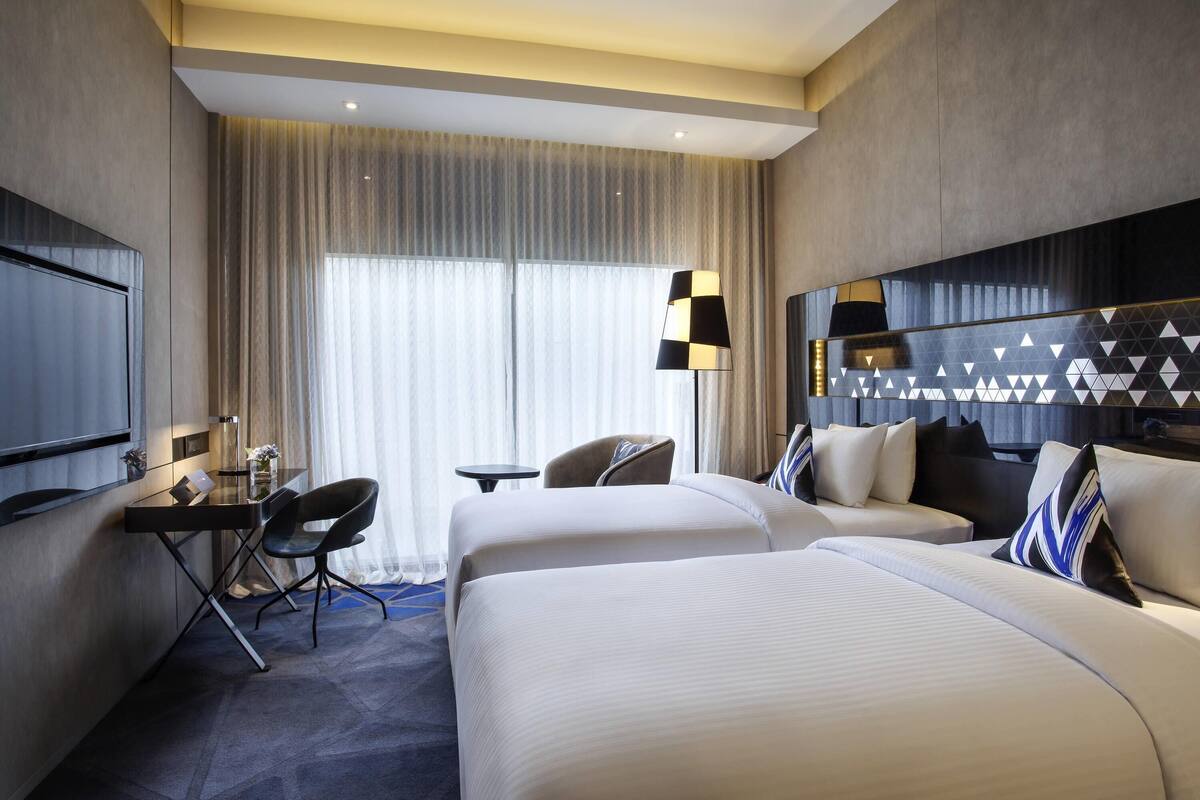 Une chambre d’hôtel avec deux lits, un bureau avec une chaise, une télévision et une grande fenêtre avec des rideaux.