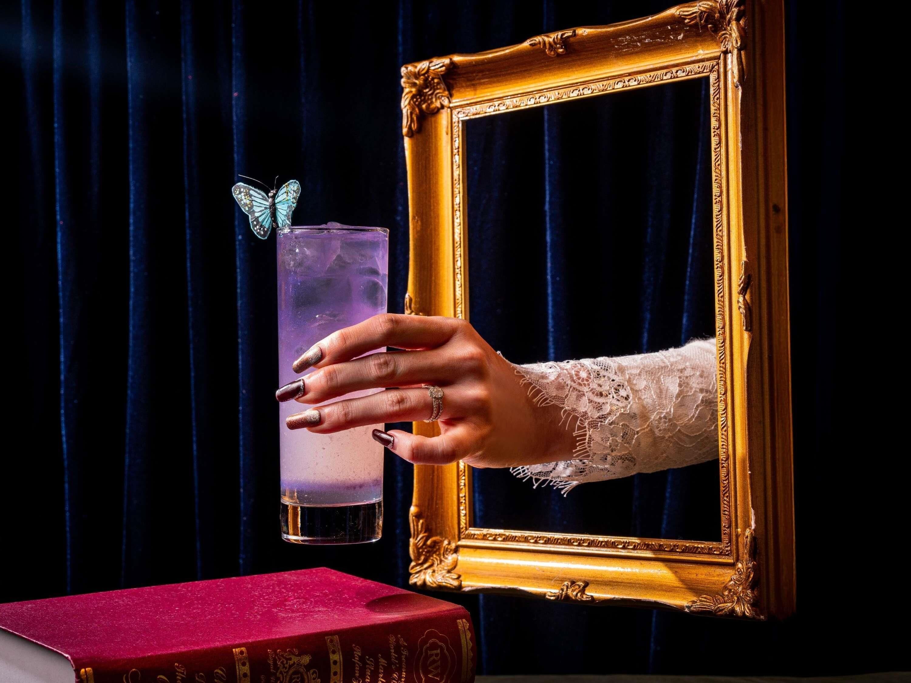 Een hand die vasthoudt een glas met een paars drankje, een boek en een spiegel met gouden lijst.