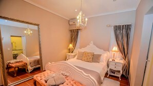 A bedroom with a large bed, a bench, a nightstand, a mirror, and a chandelier. - Lavender House (Mbombela)