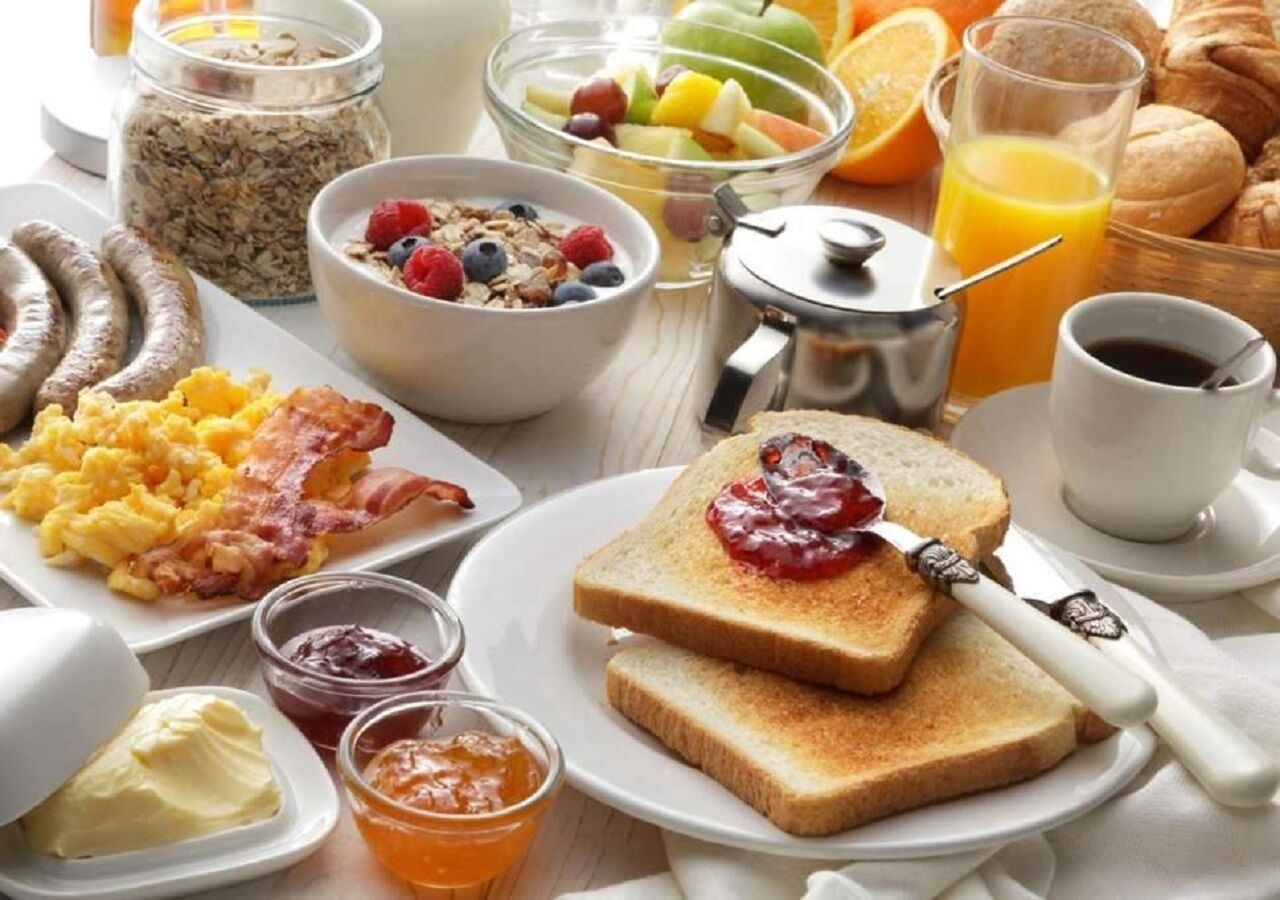 A breakfast table with various food items including scrambled eggs, bacon, toast with jam, cereal, and fruit.