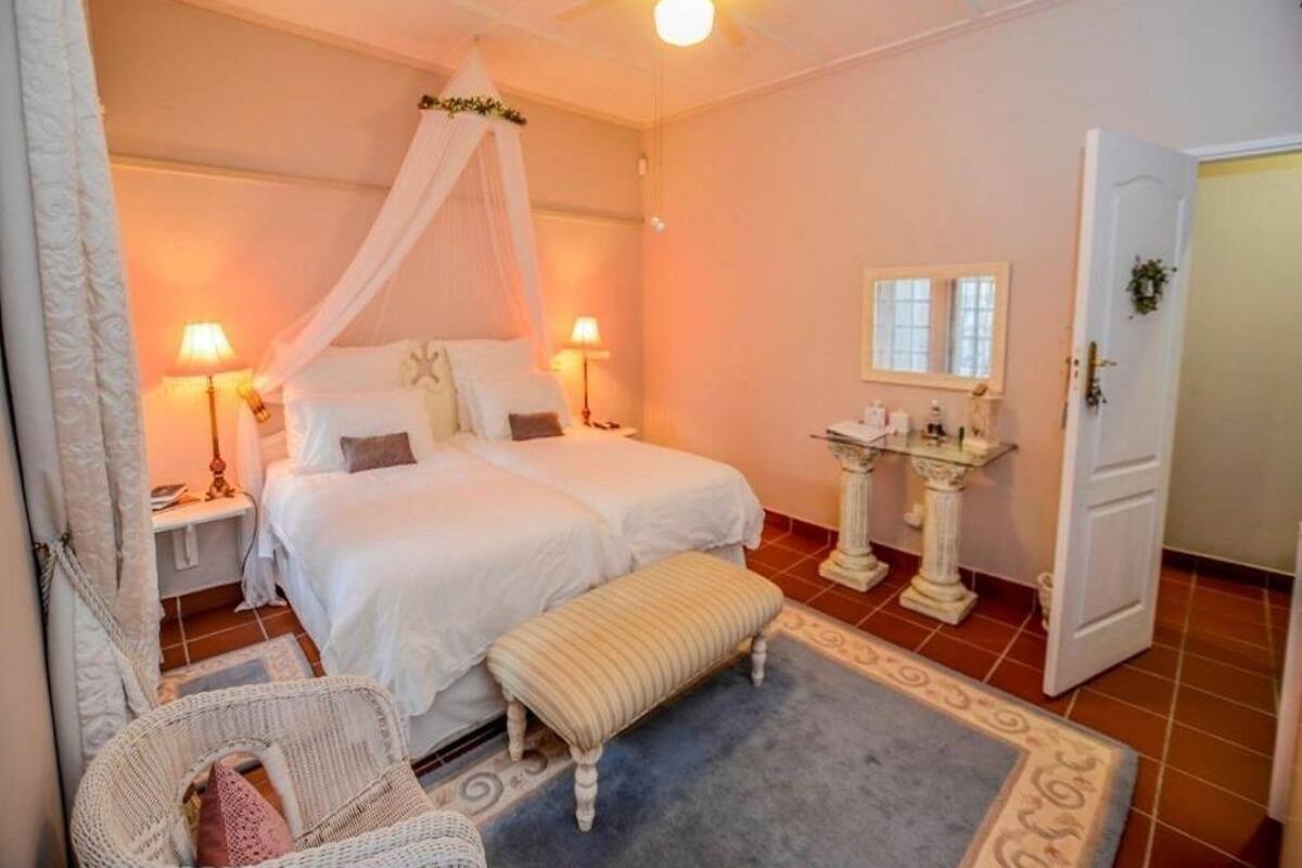 A bedroom with a canopy bed, bedside lamps, a small table, a bench, and a bathroom with a pedestal sink and mirror.