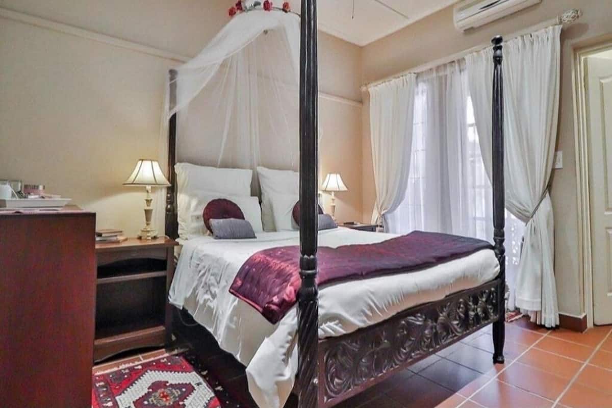A four-poster bed with white linens and a purple throw, a bedside table with a lamp, and a red patterned rug.