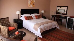 A hotel room with a bed, two bedside tables, a desk with a chair, a small refrigerator, and a wicker chair with a red cushion.