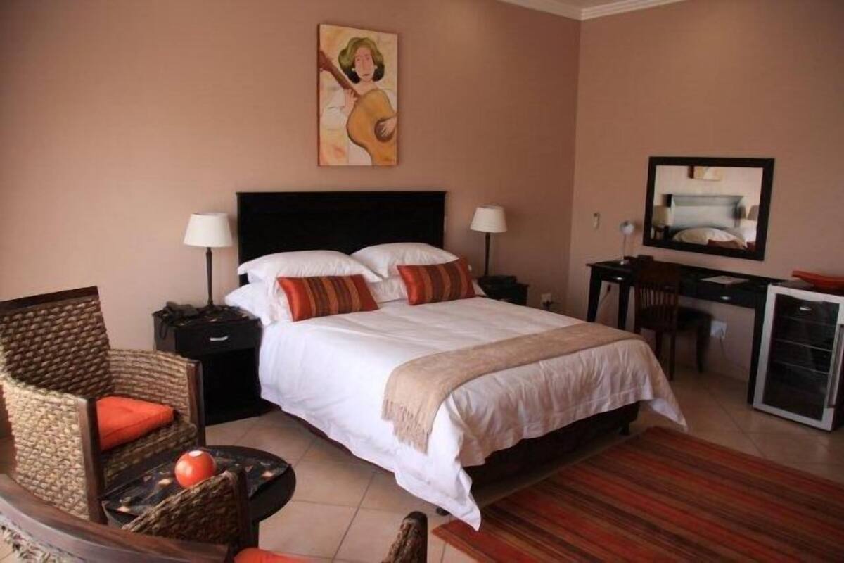 Een hotelkamer met een bed, twee nachtkastjes, een bureau met stoel, een kleine koelkast en een rieten stoel met een rode kussens.