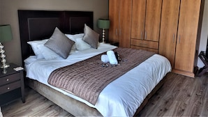 A neatly made bed with a brown quilt, two pillows, and a folded towel. There is a wooden wardrobe with multiple doors and a nightstand with a lamp. - Mandalay Beach B & B (Cape Town)