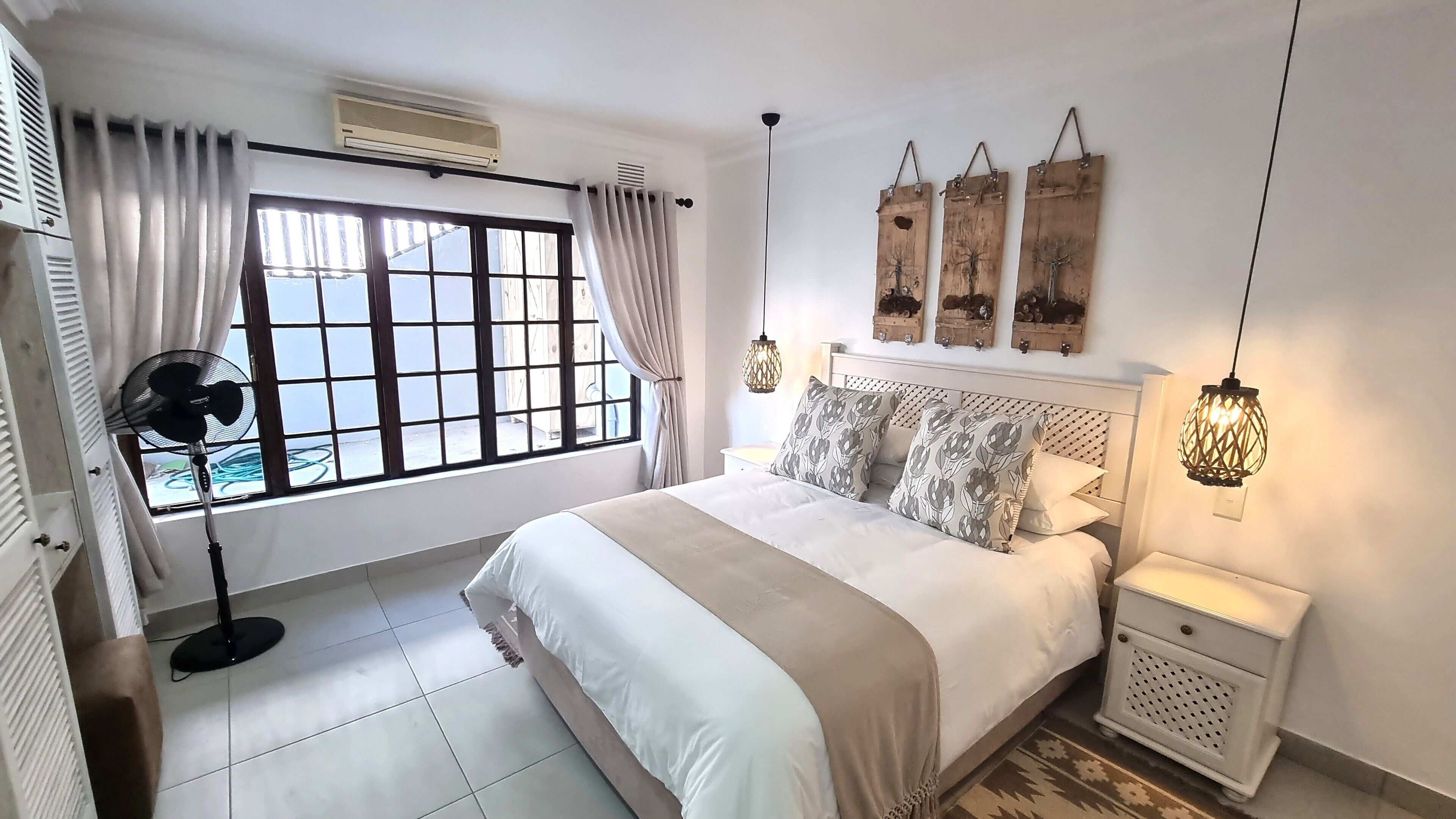 A bedroom with a large bed, a standing fan, a bedside table, and a window with curtains.
