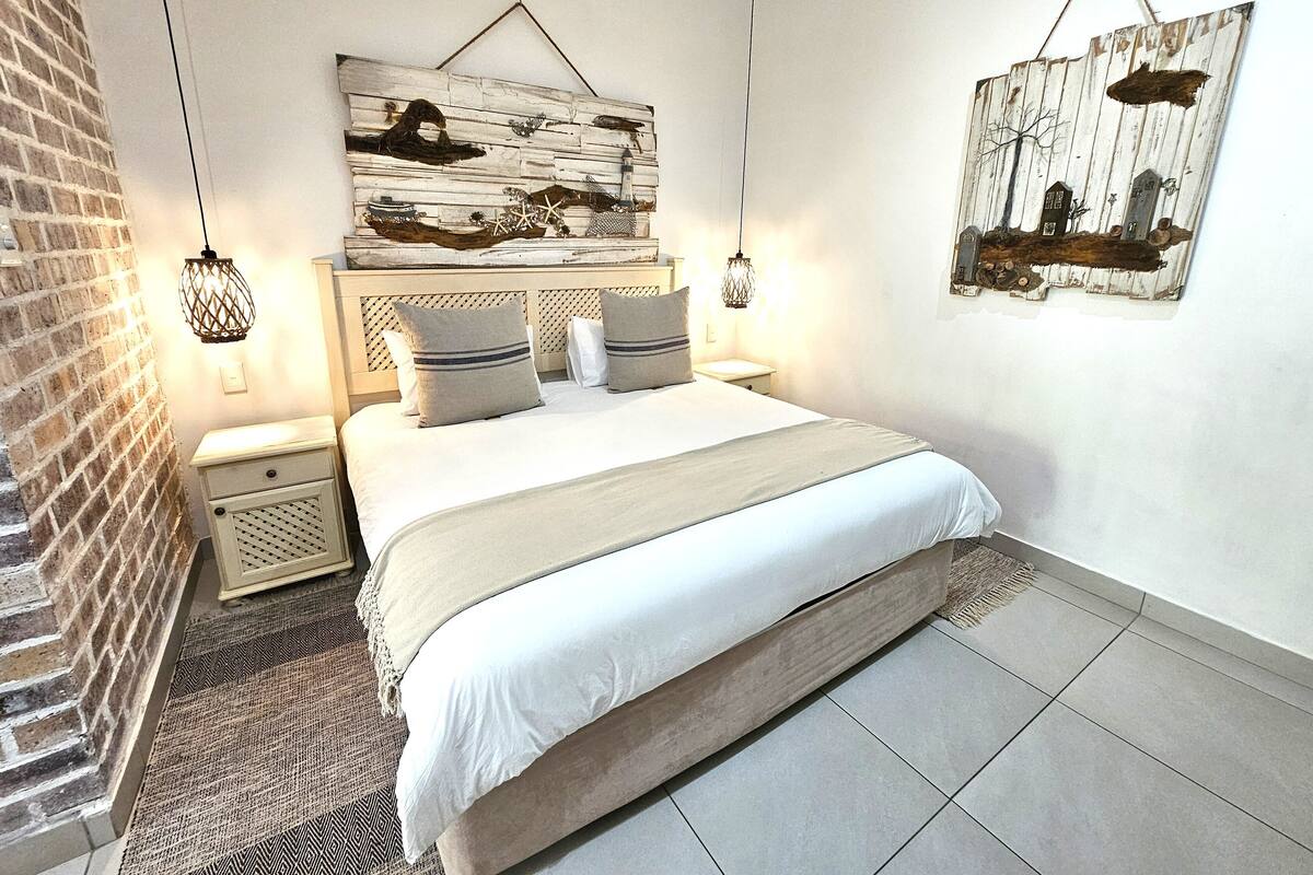 A bedroom with a bed, nightstands, a wall-mounted artwork, and a hanging light fixture.
