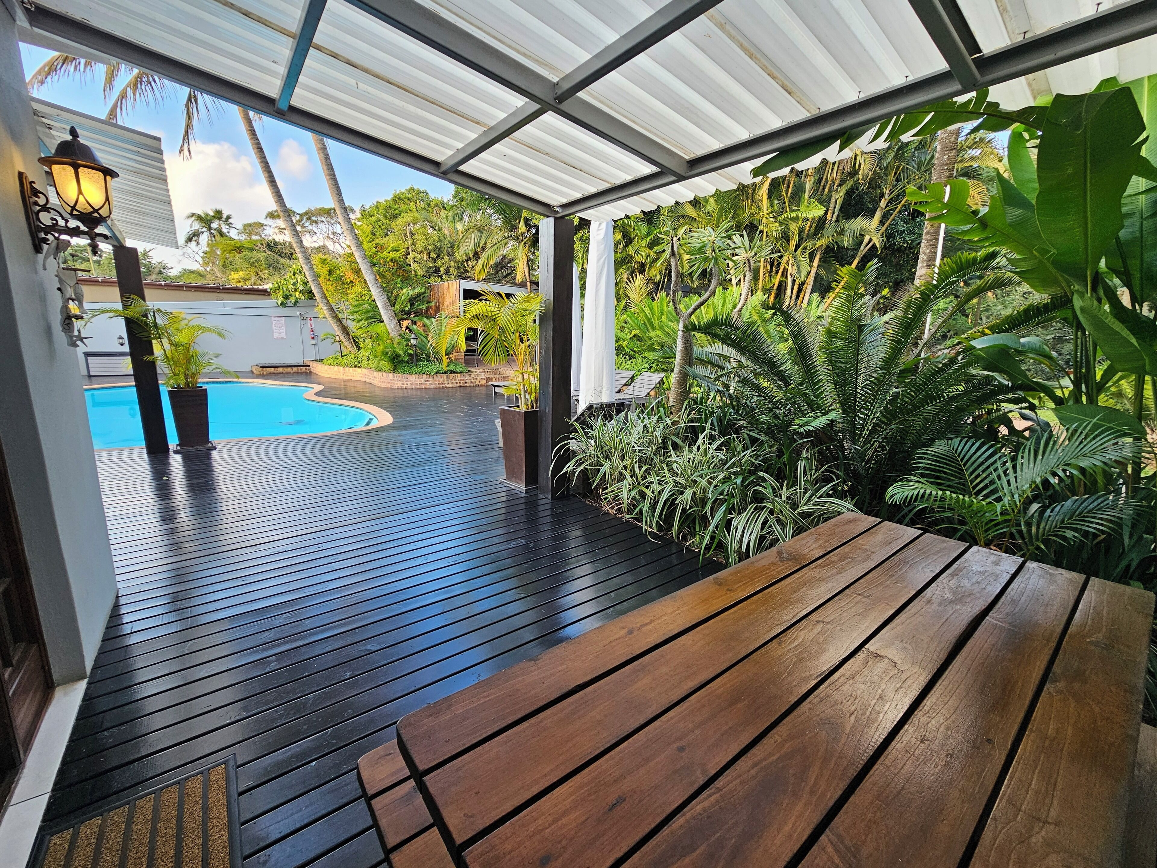 A covered outdoor area with a wooden deck, a pool, and lush tropical plants.