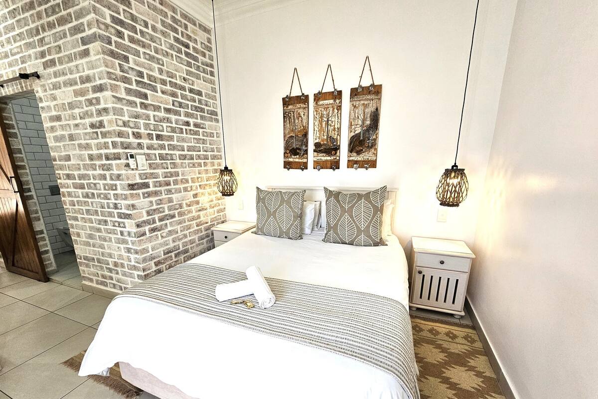 A hotel room with a brick wall, a bed with patterned bedding, a nightstand, and wall-mounted artwork.