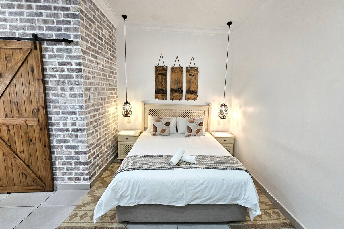 A bedroom with a brick wall, a wooden door, a bed with white bedding, two bedside tables, and wall-mounted lamps.
