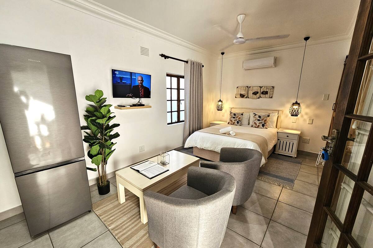 A modern hotel room with a bed, two armchairs, a desk, a flat-screen TV, a refrigerator, and a potted plant.