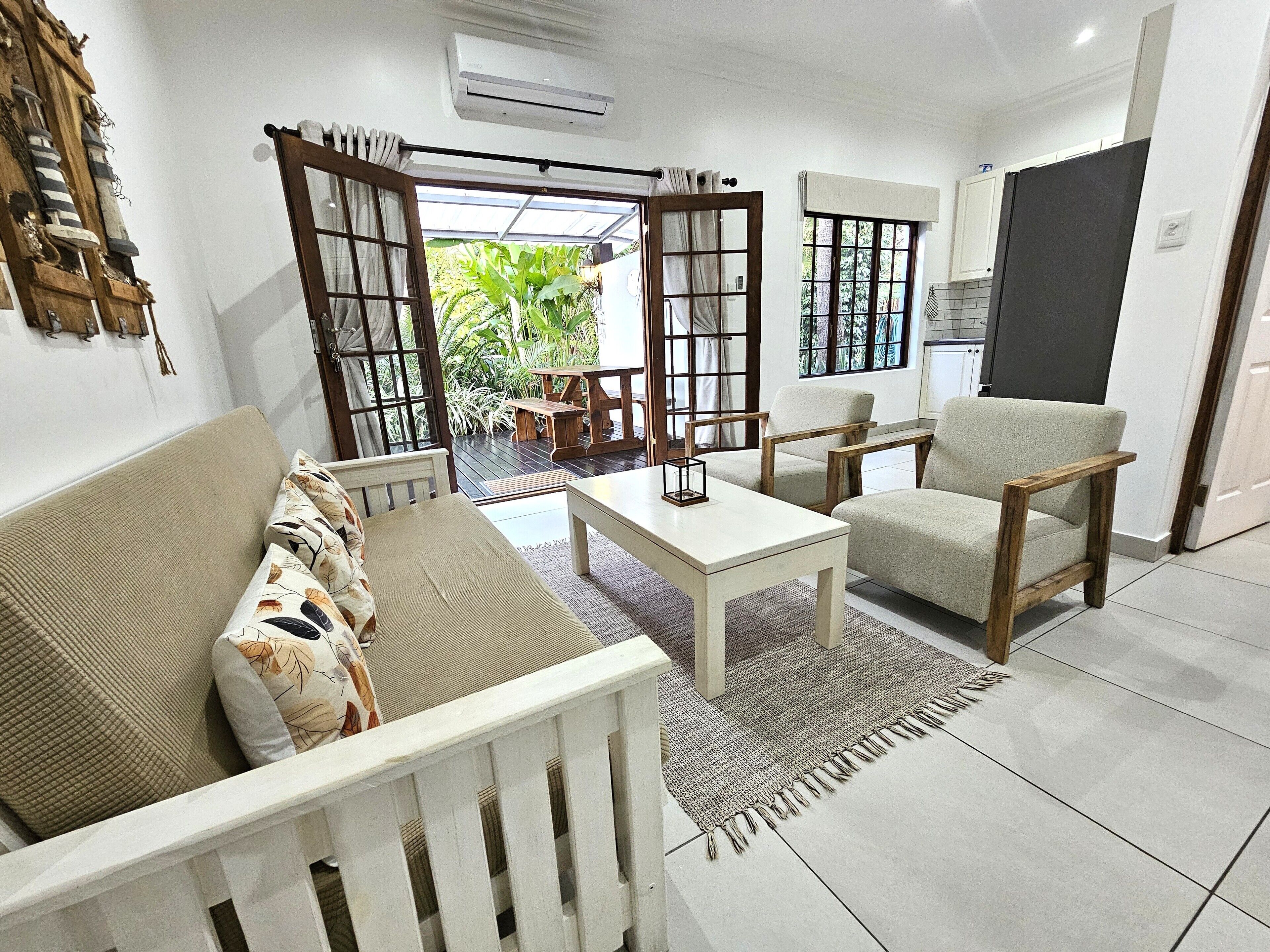 A modern living room with a sofa, armchair, coffee table, and a view of a garden through open doors.