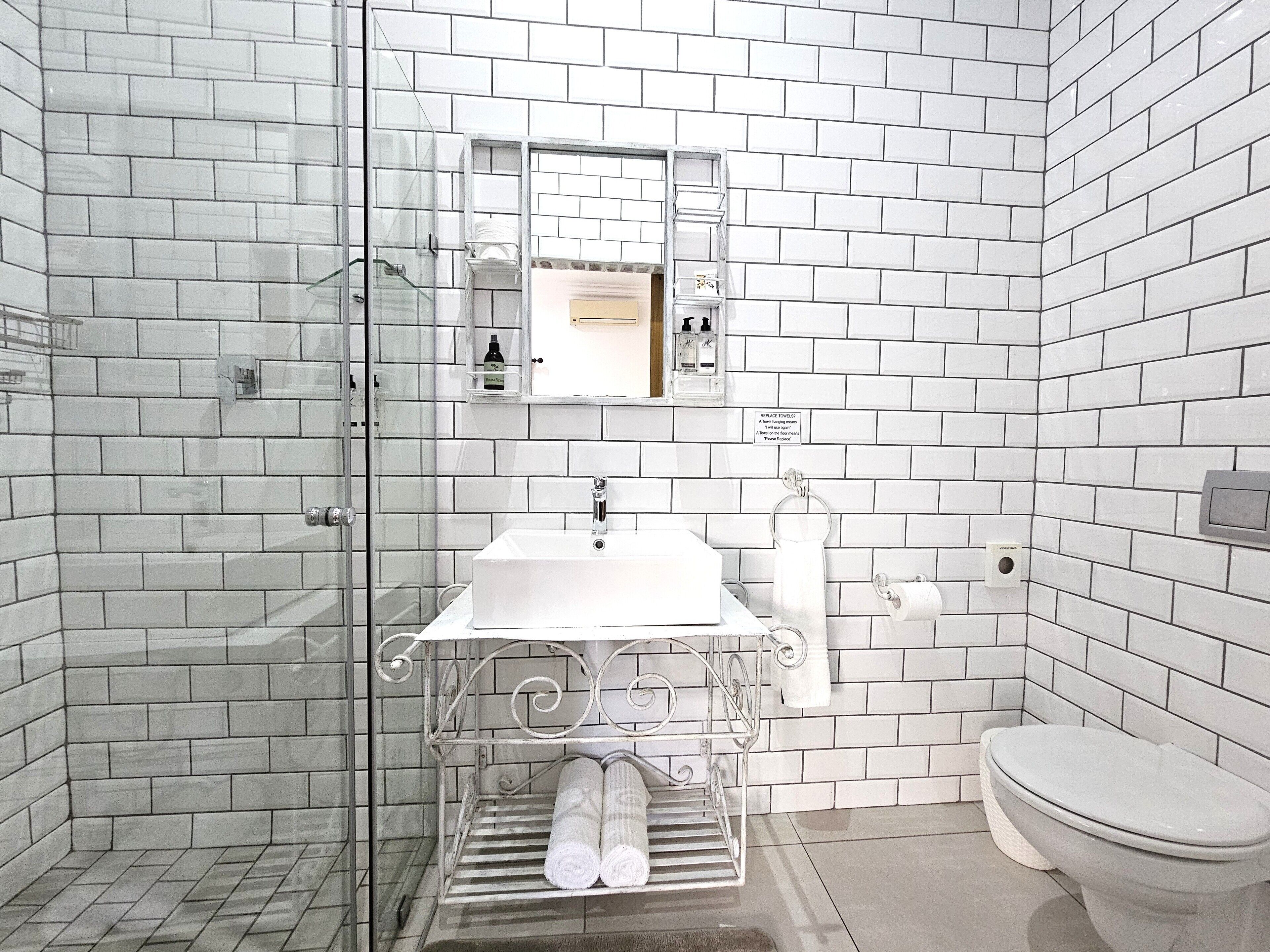 Un baño con paredes de azulejos blancos, cabina de ducha de vidrio, lavamanos blanco con espejo sobre él y un inodoro.