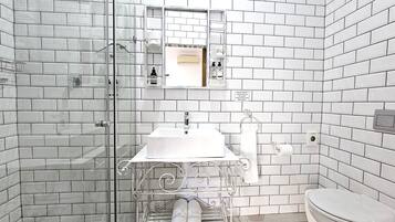 Un baño con paredes de azulejos blancos, cabina de ducha de vidrio, lavamanos blanco con espejo sobre él y un inodoro.