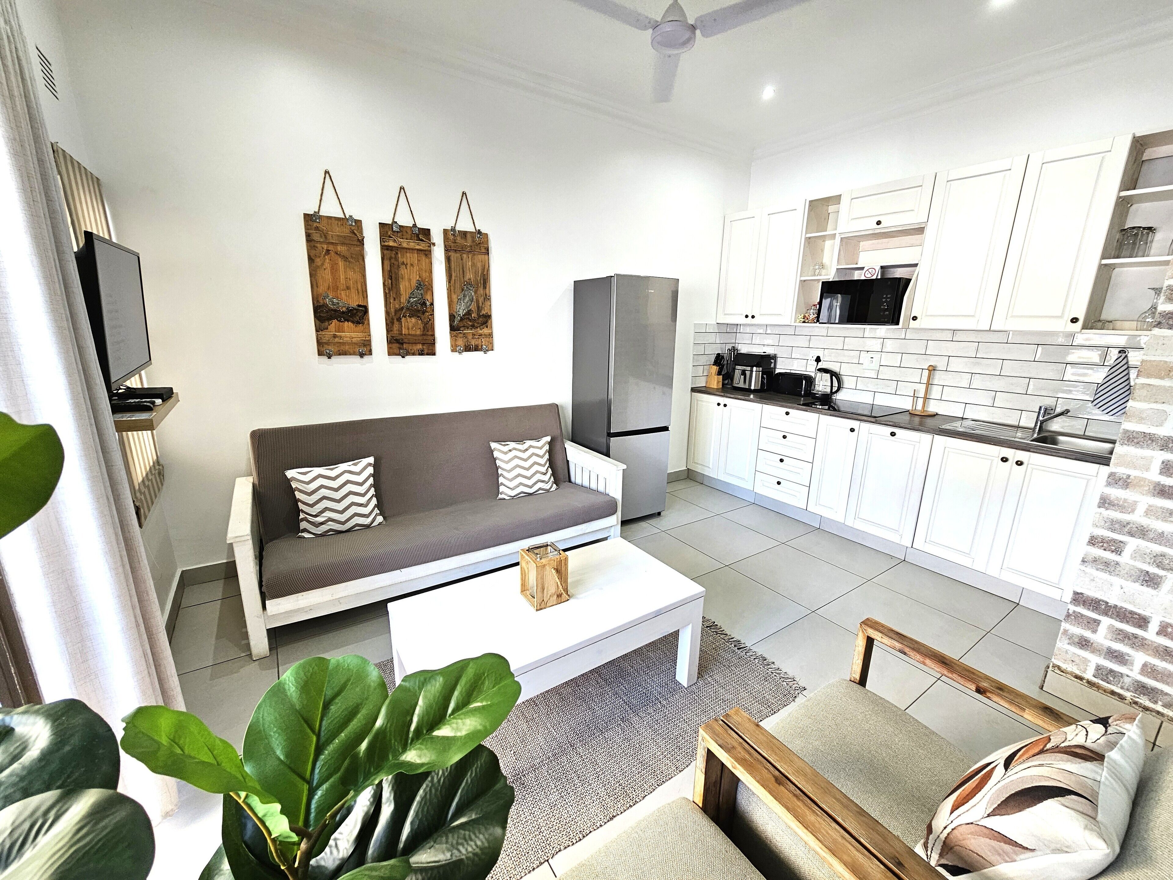 A modern living room with a sofa, coffee table, and a TV. Adjacent is a kitchen with white cabinets and a brick accent wall.