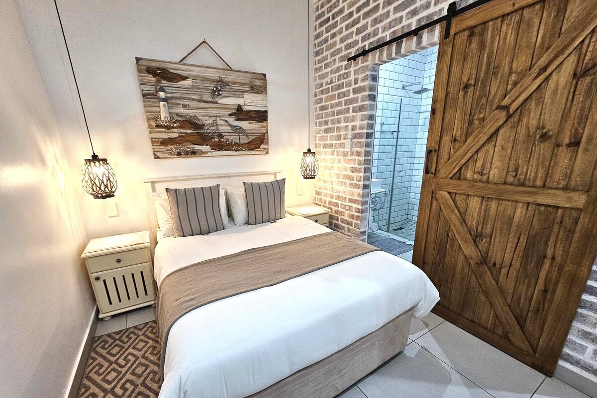 A bedroom with a large bed, a wooden door, a brick wall, and a hanging artwork.