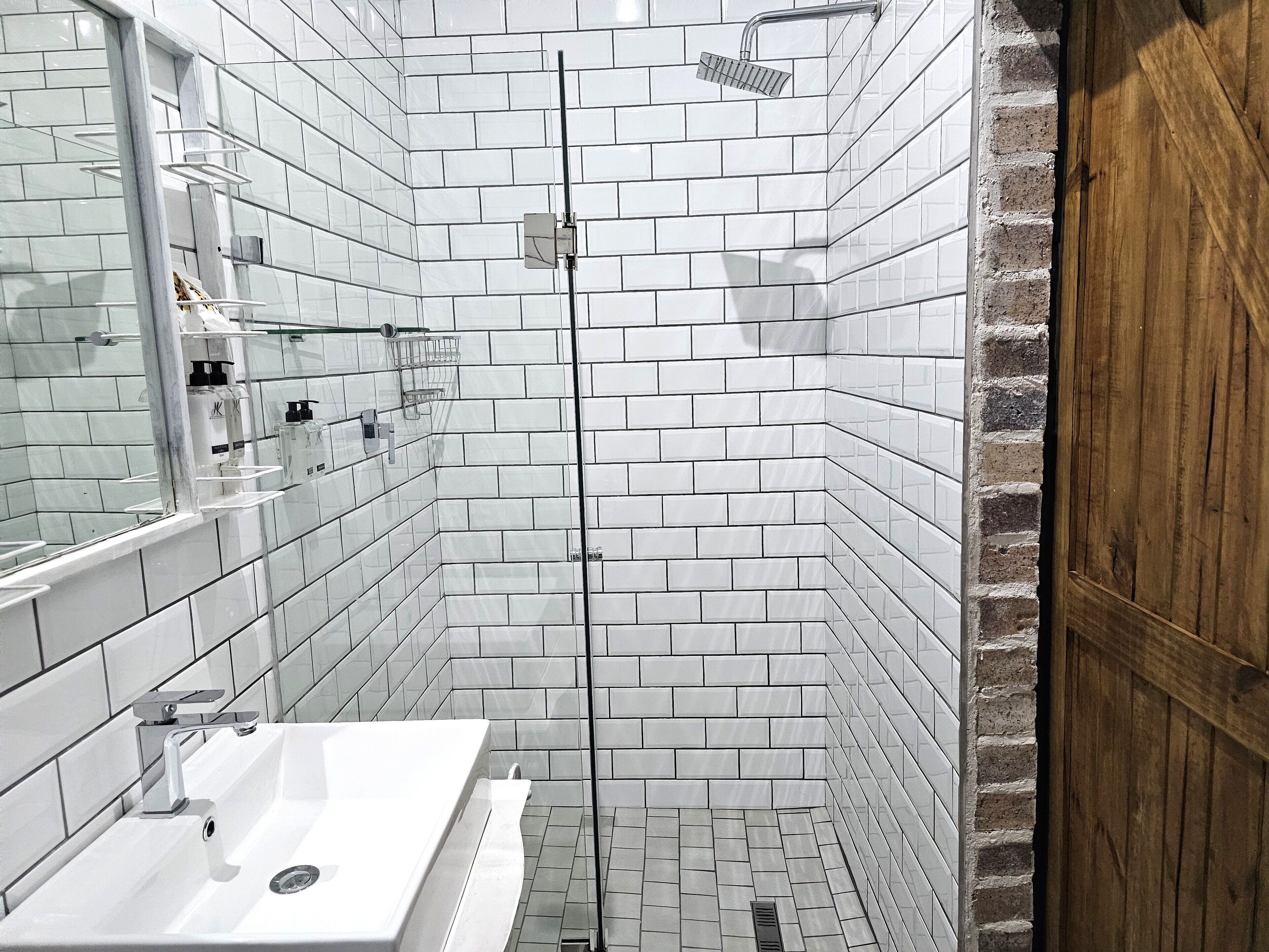 Ein modernes Badezimmer mit weißen, in Ziegeloptik angeordneten Fliesen, einer Glasdusche und einer Holztür.