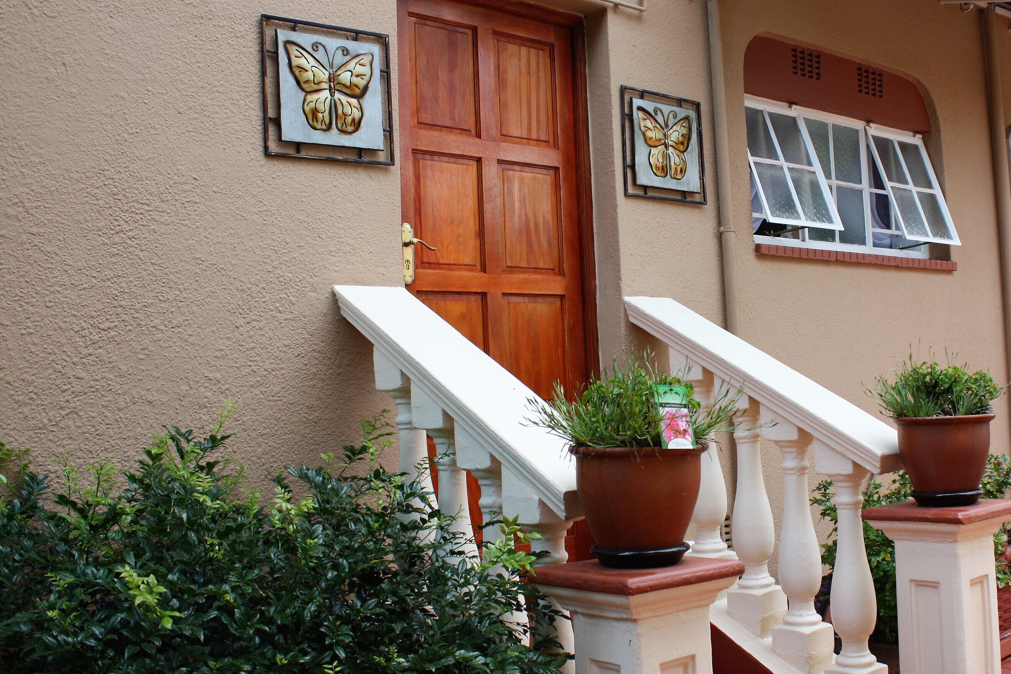 A house with a wooden door, a window with shutters, and a staircase with white balusters. There are potted plants on the railing.
