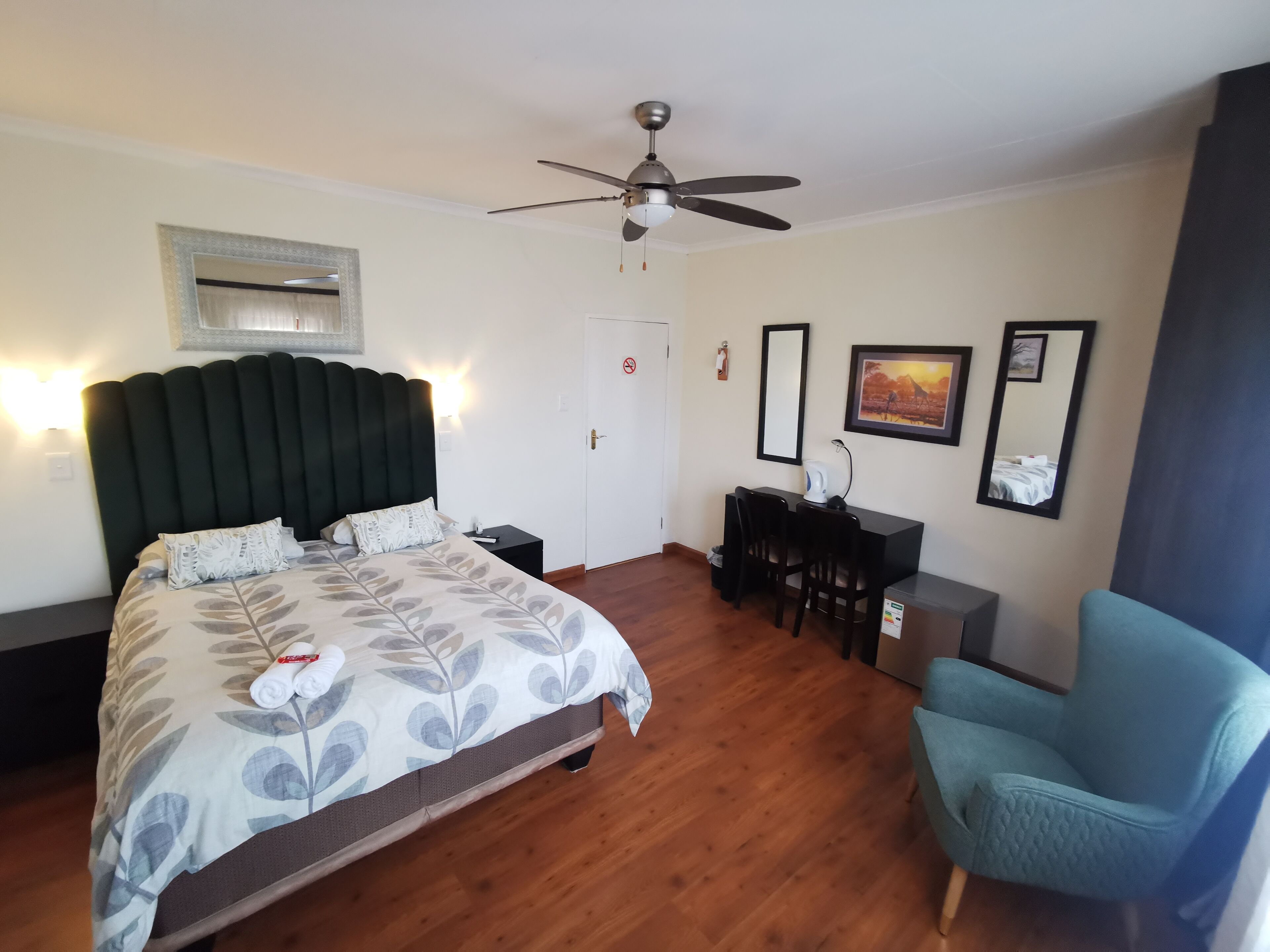 Habitación de hotel con cama, escritorio, silla, ventilador de techo y obras de arte en las paredes.
