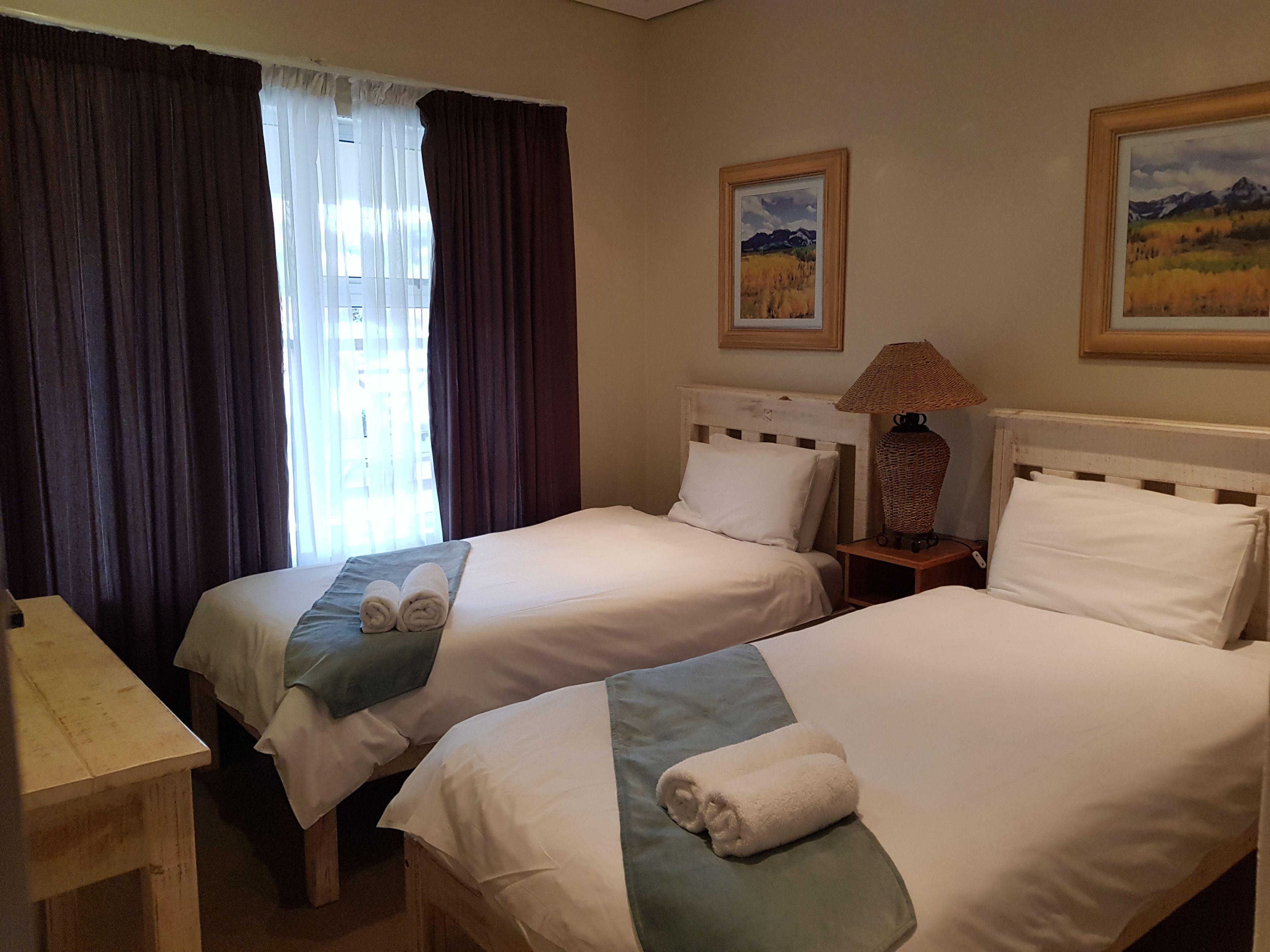 A hotel room with two beds, a desk, a lamp, and two framed pictures on the wall.