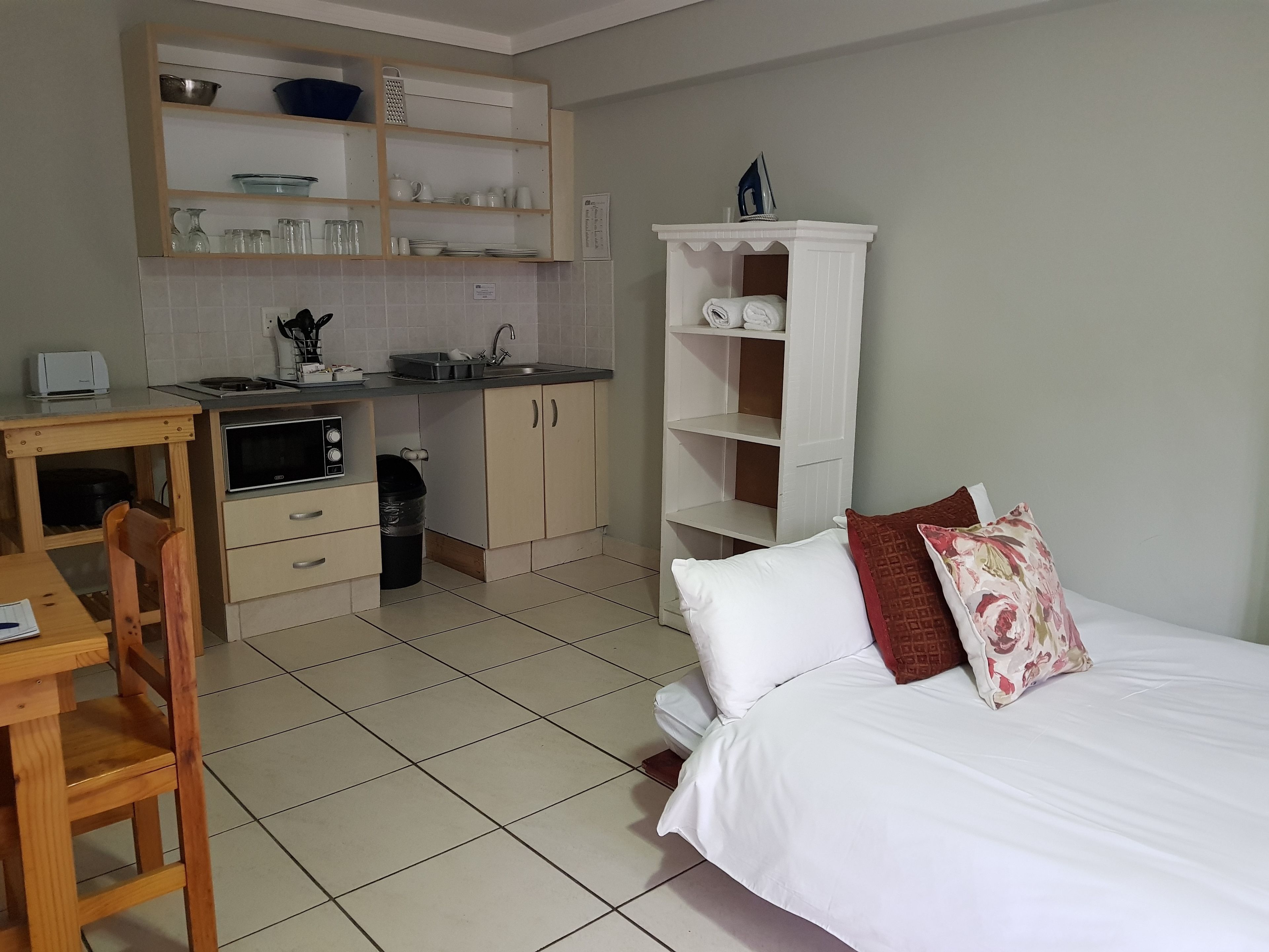 A compact hotel room with a kitchenette, a bed with white and floral pillows, and a wooden chair.