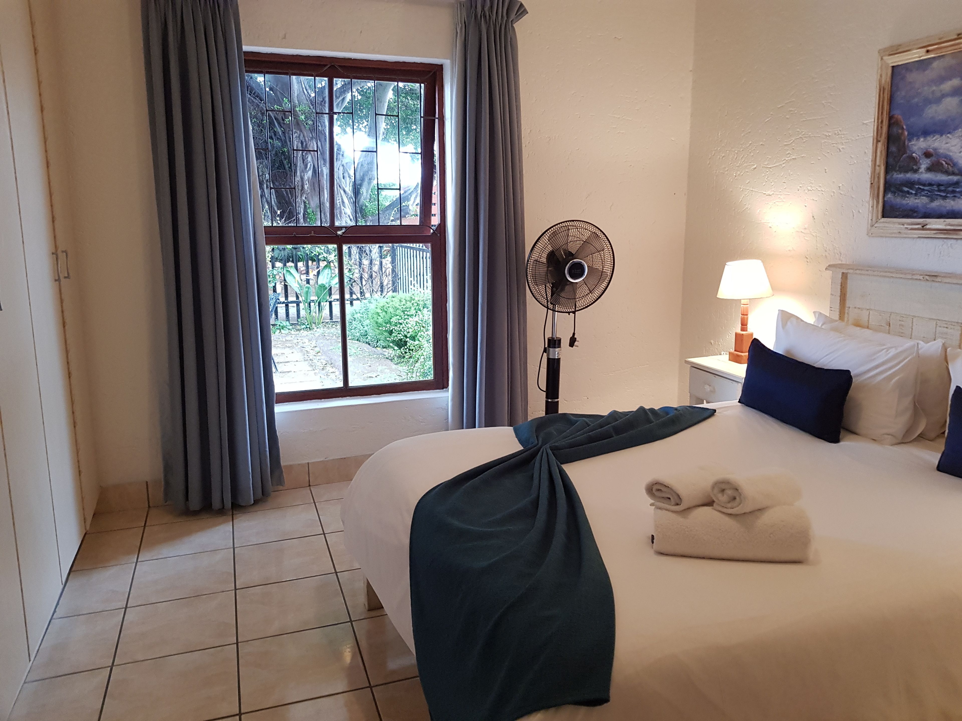 A hotel room with a bed, a window with curtains, a standing fan, a bedside lamp, and a painting on the wall.