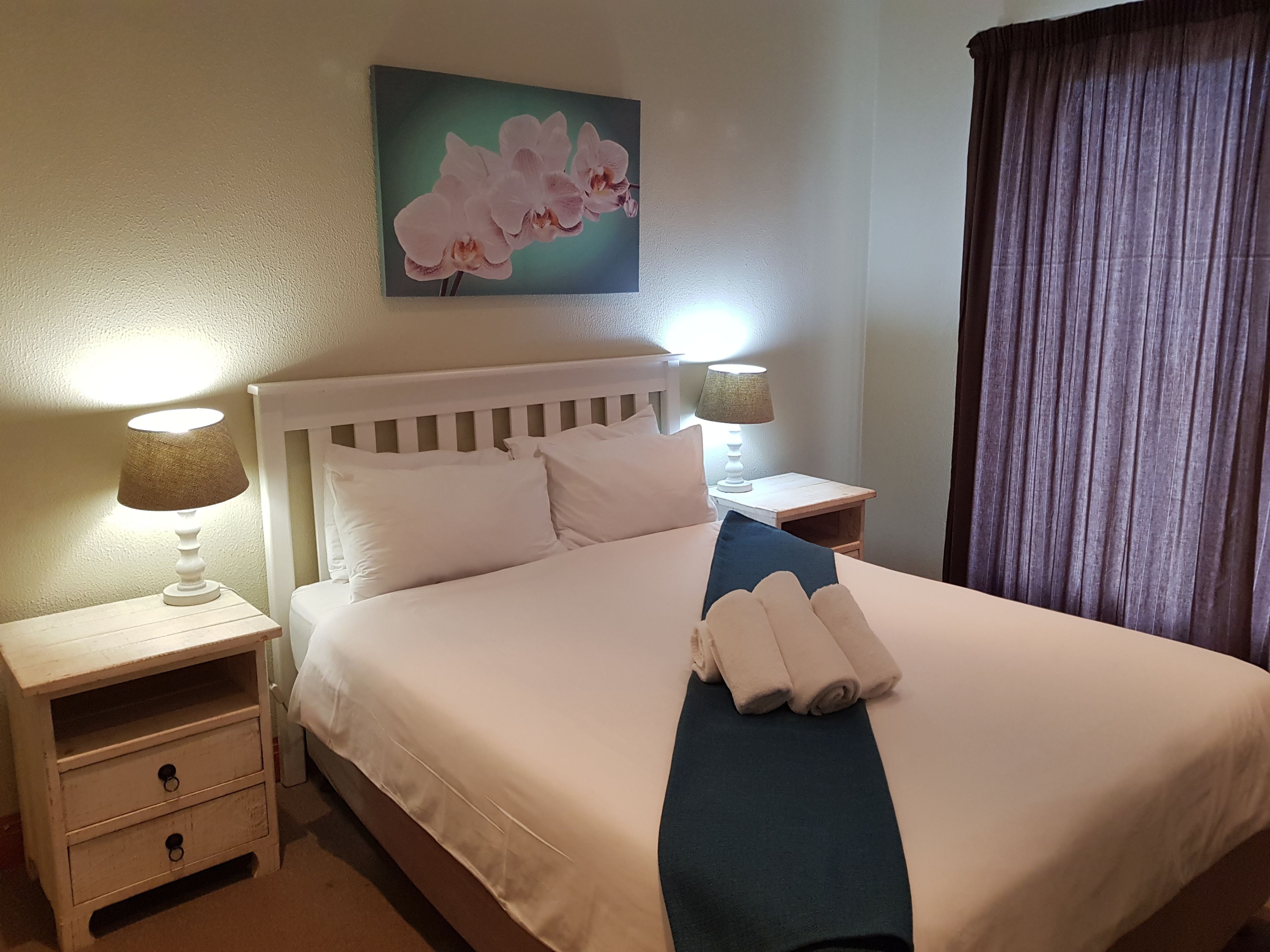 Un lit bien fait, avec du linge de lit blanc, deux tables de chevet avec des lampes, un tableau représentant des orchidées accroché au mur et un rideau foncé à la fenêtre.