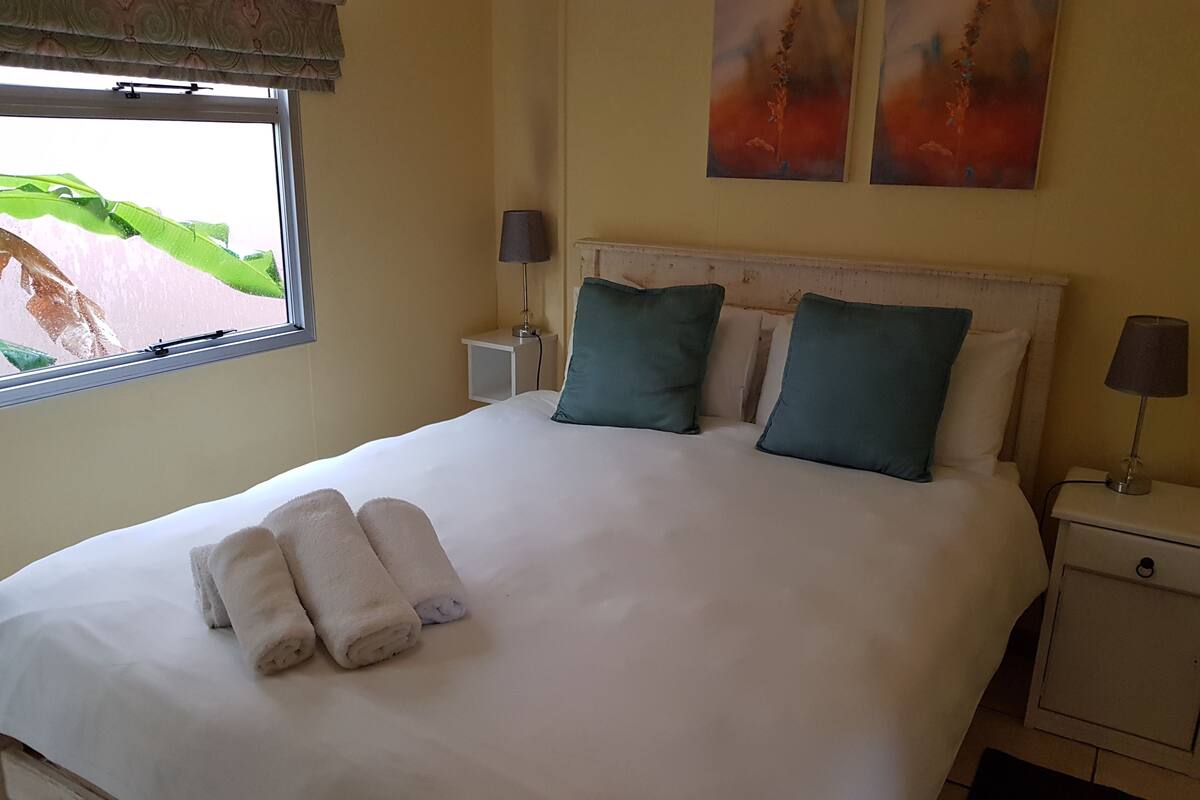 A neatly made bed with white linens and two rolled towels, a bedside table with a lamp, and a window with a view of greenery.