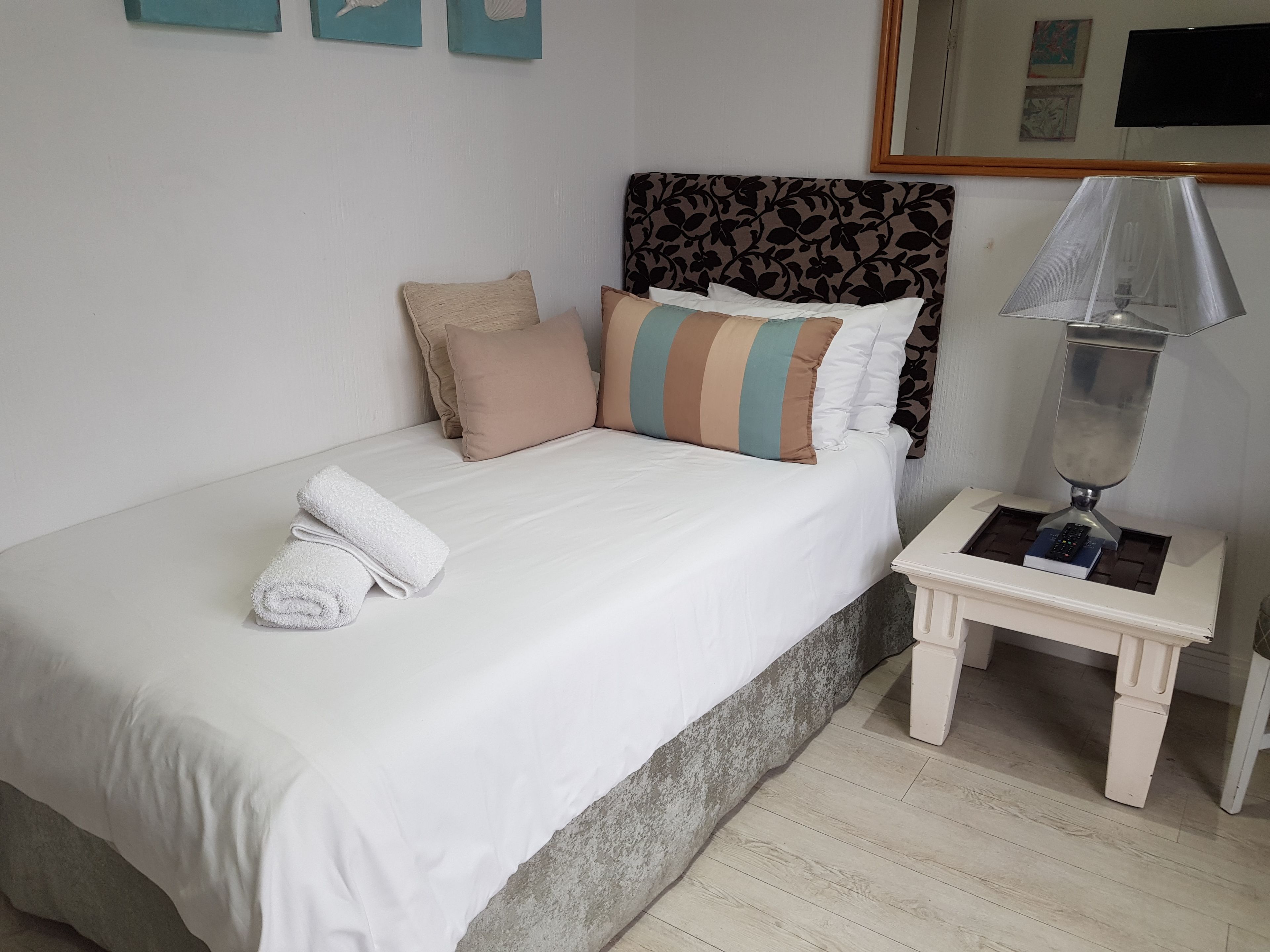 Una cama bien hecha con edredón blanco, un almohadón a rayas y un almohadón color beige. Se observa una mesita de noche con una lámpara y un libro.