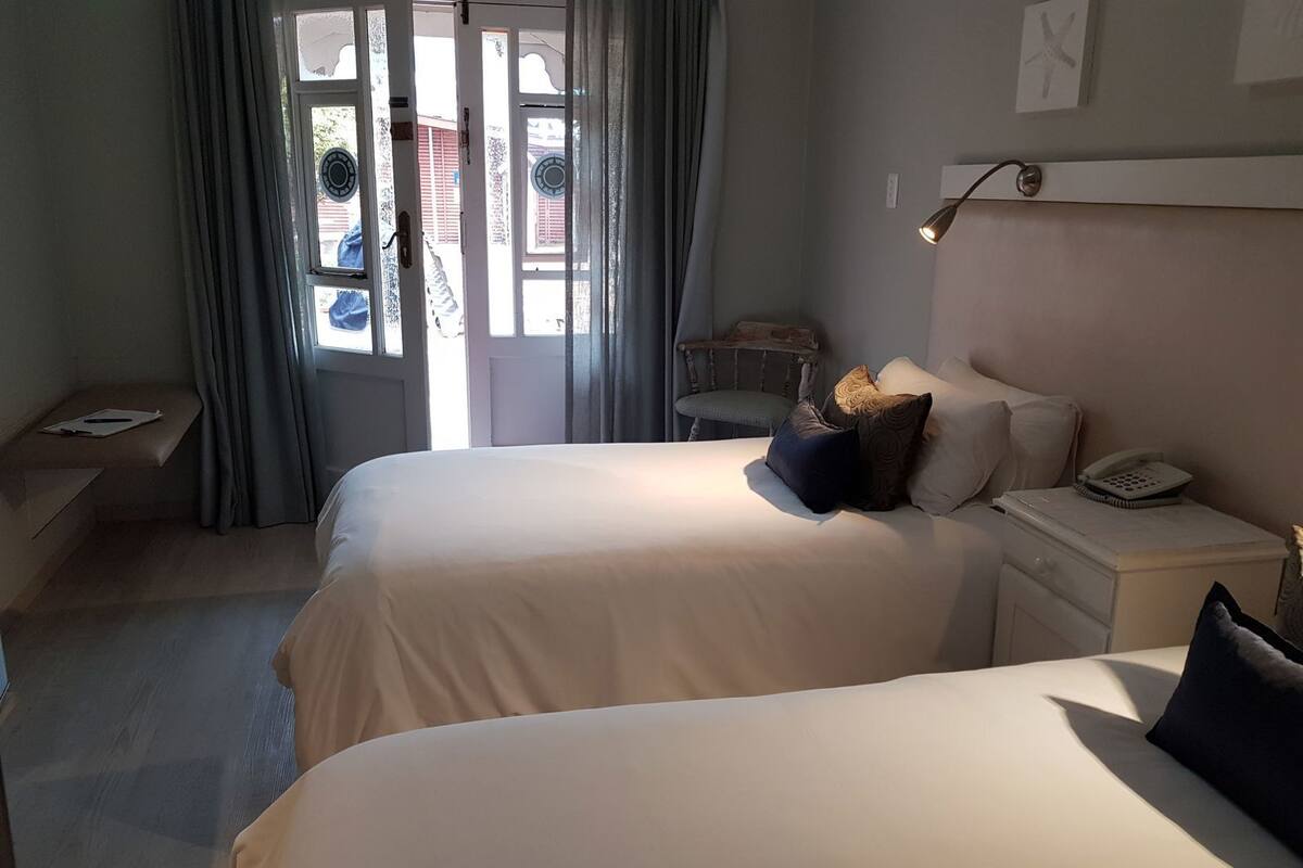 A hotel room with two beds, a desk, a chair, a bedside table with a lamp, and a window with curtains.