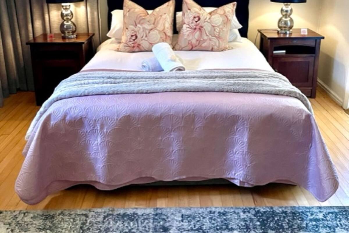 A neatly made bed with a pink bedspread and matching pillows, flanked by two bedside tables with lamps, in a room with wooden flooring and a patterned rug.