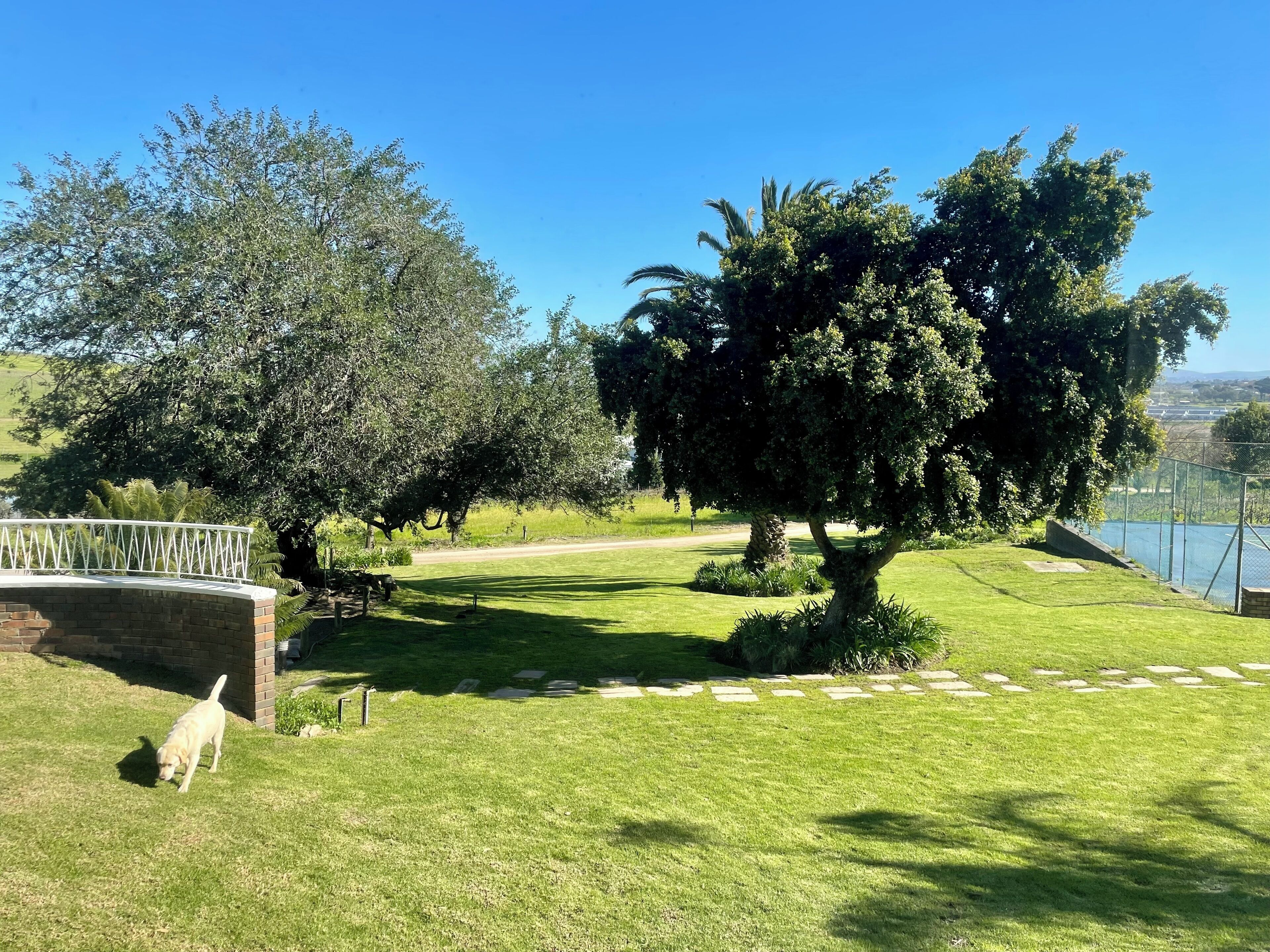 A well-maintained grassy area with a brick structure, a white fence, and a large tree providing shade.
