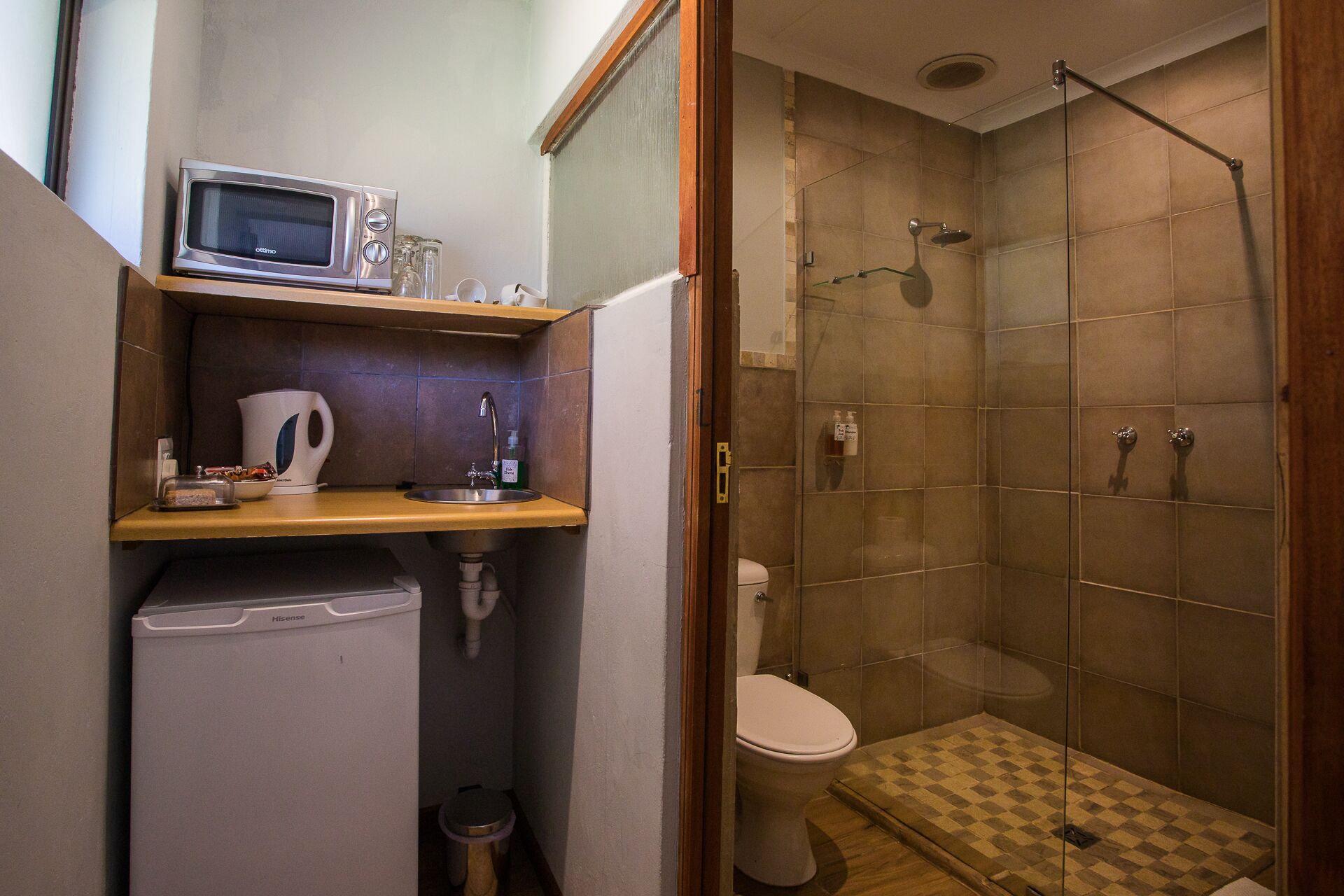 Un baño pequeño con inodoro, ducha con puerta de vidrio, lavamanos y un microondas encima, además de un pequeño espacio de almacenamiento.