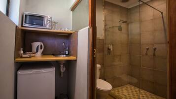Un baño pequeño con inodoro, ducha con puerta de vidrio, lavamanos y un microondas encima, además de un pequeño espacio de almacenamiento.