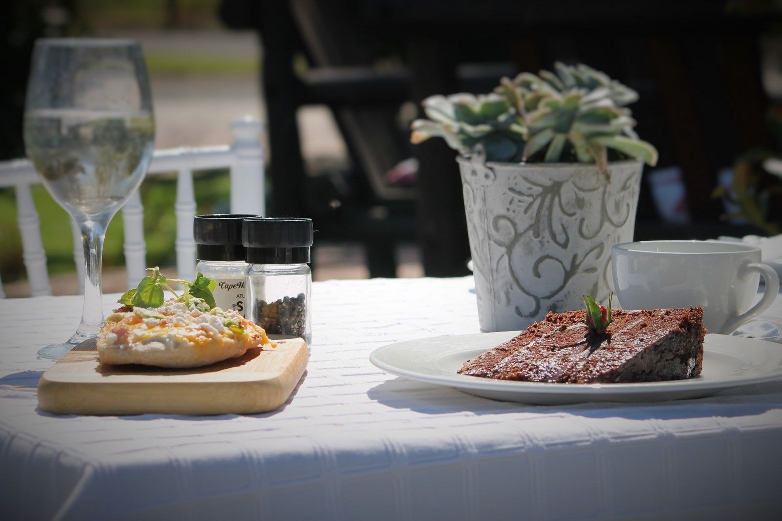 A table with a slice of cake, a glass of white wine, and a small dish of food, along with salt and pepper shakers and a potted plant.