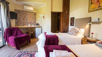 Une chambre d’hôtel comprenant un lit, deux tables de chevet, un bureau avec une chaise, un petit réfrigérateur, une fenêtre avec des rideaux et un tableau au mur.