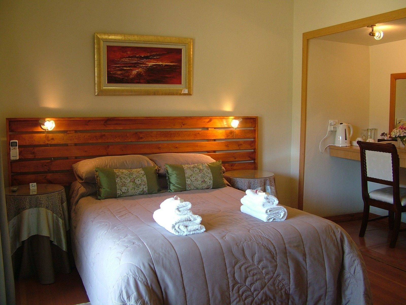 A neatly made bed with a wooden headboard, a painting above it, and a small table with a kettle and flowers beside the bed.