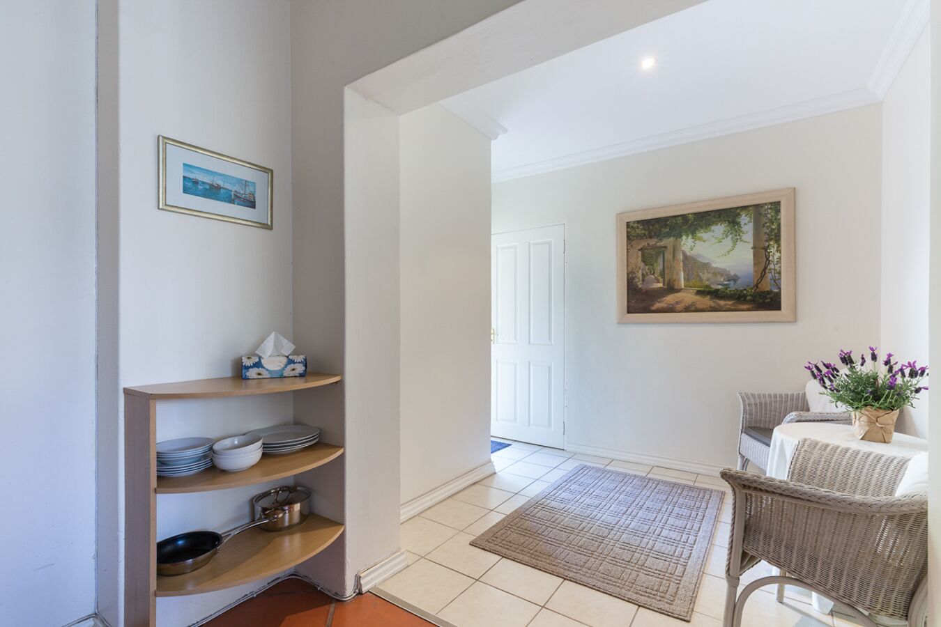 A hallway with a wooden shelf holding plates and bowls, a framed painting on the wall, and a wicker chair with a table and vase.