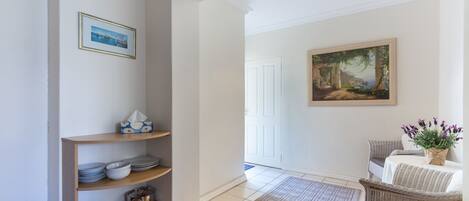 A hallway with a wooden shelf holding plates and bowls, a framed painting on the wall, and a wicker chair with a table and vase.