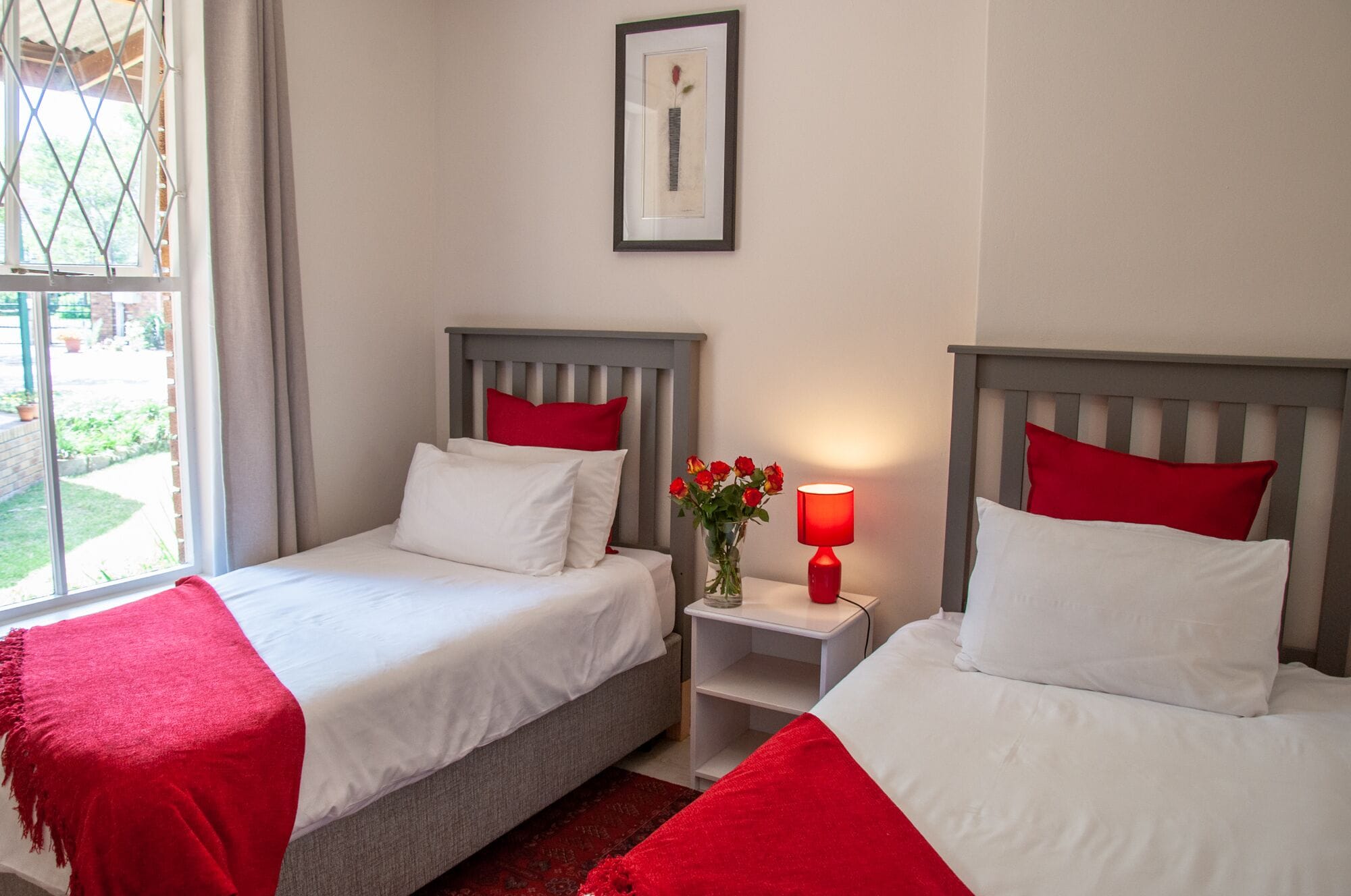 Une chambre d’hôtel avec deux lits, chacun agrémenté de draps blancs et d’oreillers rouges, une petite table de chevet avec une lampe rouge et un tableau encadré au mur.